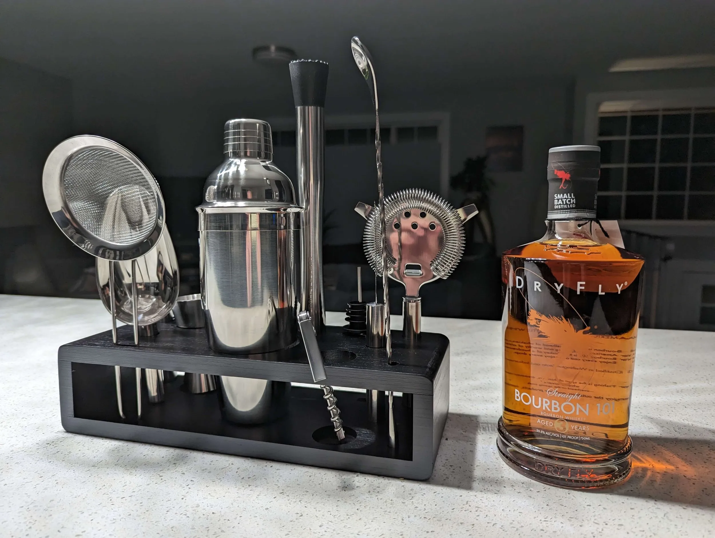 Stainless-steel cocktail tools arranged in the dark bamboo stand beside a bottle of bourbon.